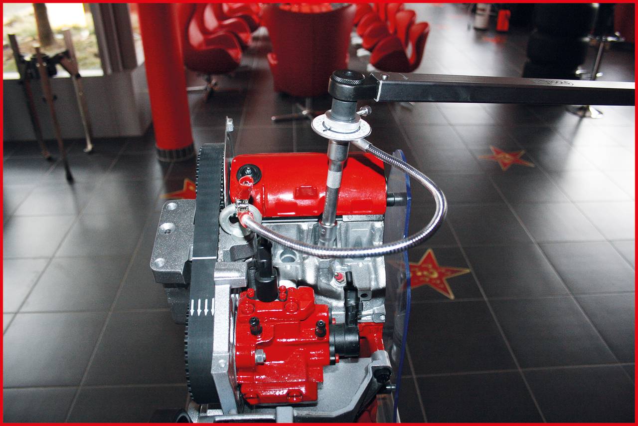 Ein roter Motorblock mit einem Drehmomentschlüssel auf einer Werkstattfläche, umgeben von roten Stühlen und schwarzen Fliesen.
