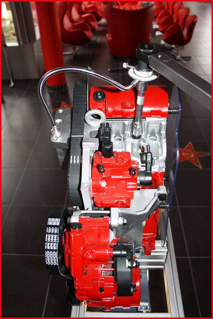 Ein roter Motor, ausgestellt in einem modernen Raum mit schwarzen Fliesen und roten Stühlen, verbindet mechanische Komponenten.