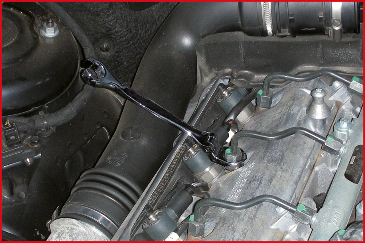 Nahaufnahme eines Motorraums mit einem Schraubenschlüssel, der an einem Bauteil befestigt ist. Links sind Schläuche und Anschlüsse erkennbar.