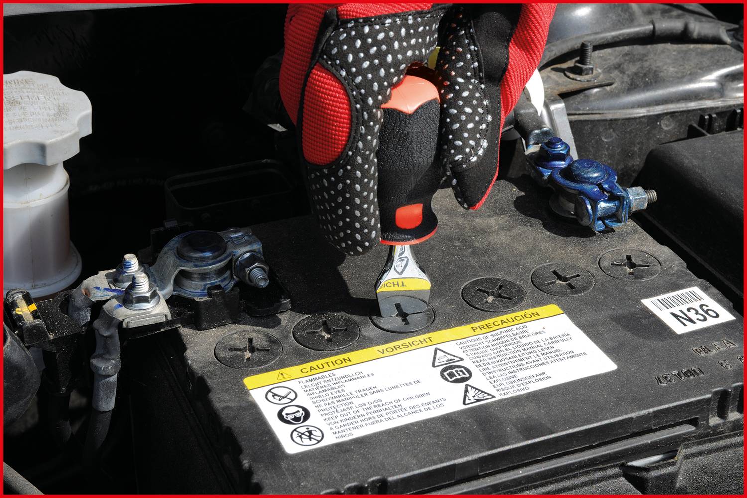 Eine Hand mit Handschuhen zieht eine Batterieabdeckung im Motorraum eines Autos ab. Die Batterie hat Warnhinweise auf der Oberseite.