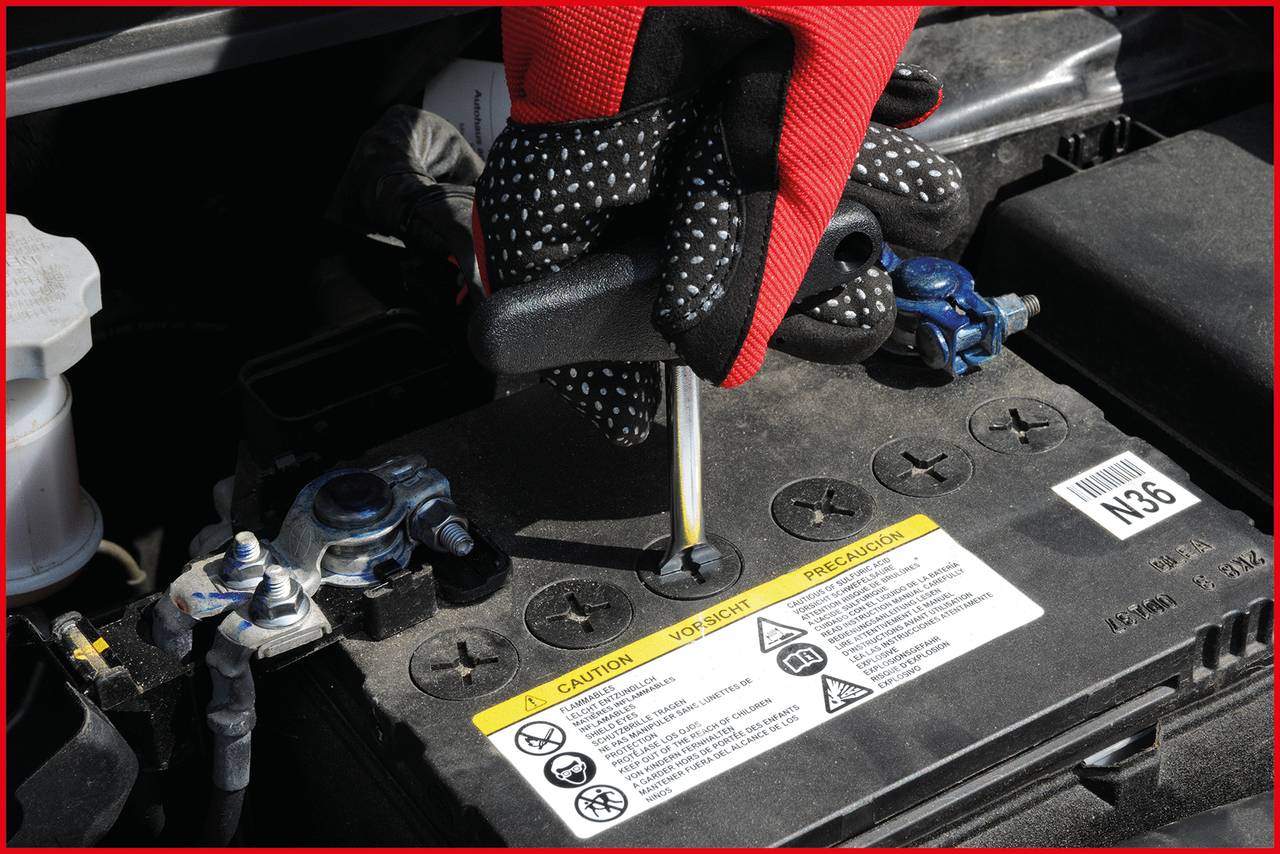 Eine Hand mit Handschuh hält einen Schraubendreher und überprüft den Flüssigkeitsstand in einer Autobatterie.