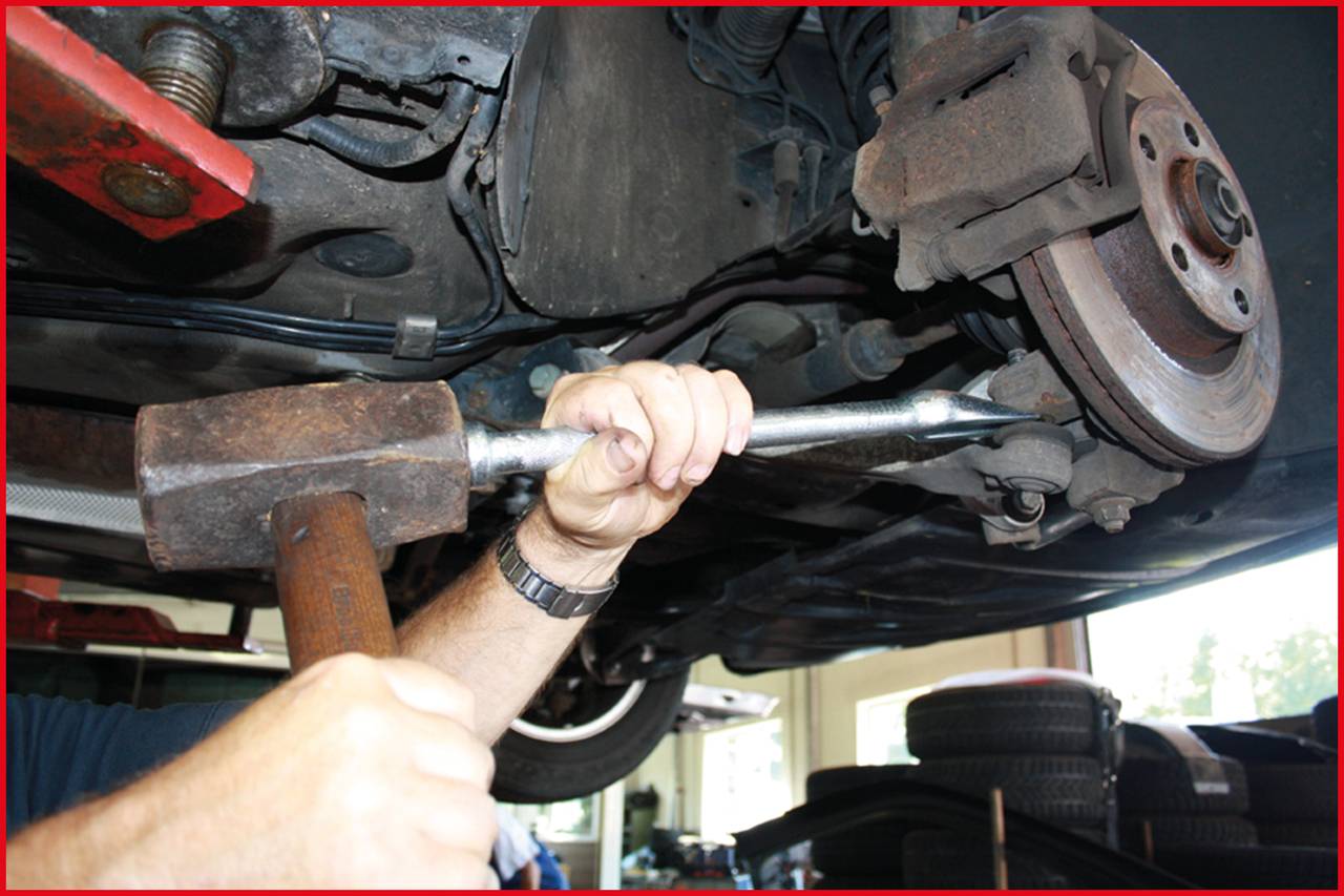 'Nahaufnahme eines Mechanikers, der mit einem Hammer an einem Auto arbeitet. Die Aufnahme zeigt den Unterboden und ein Rad des Fahrzeugs.'