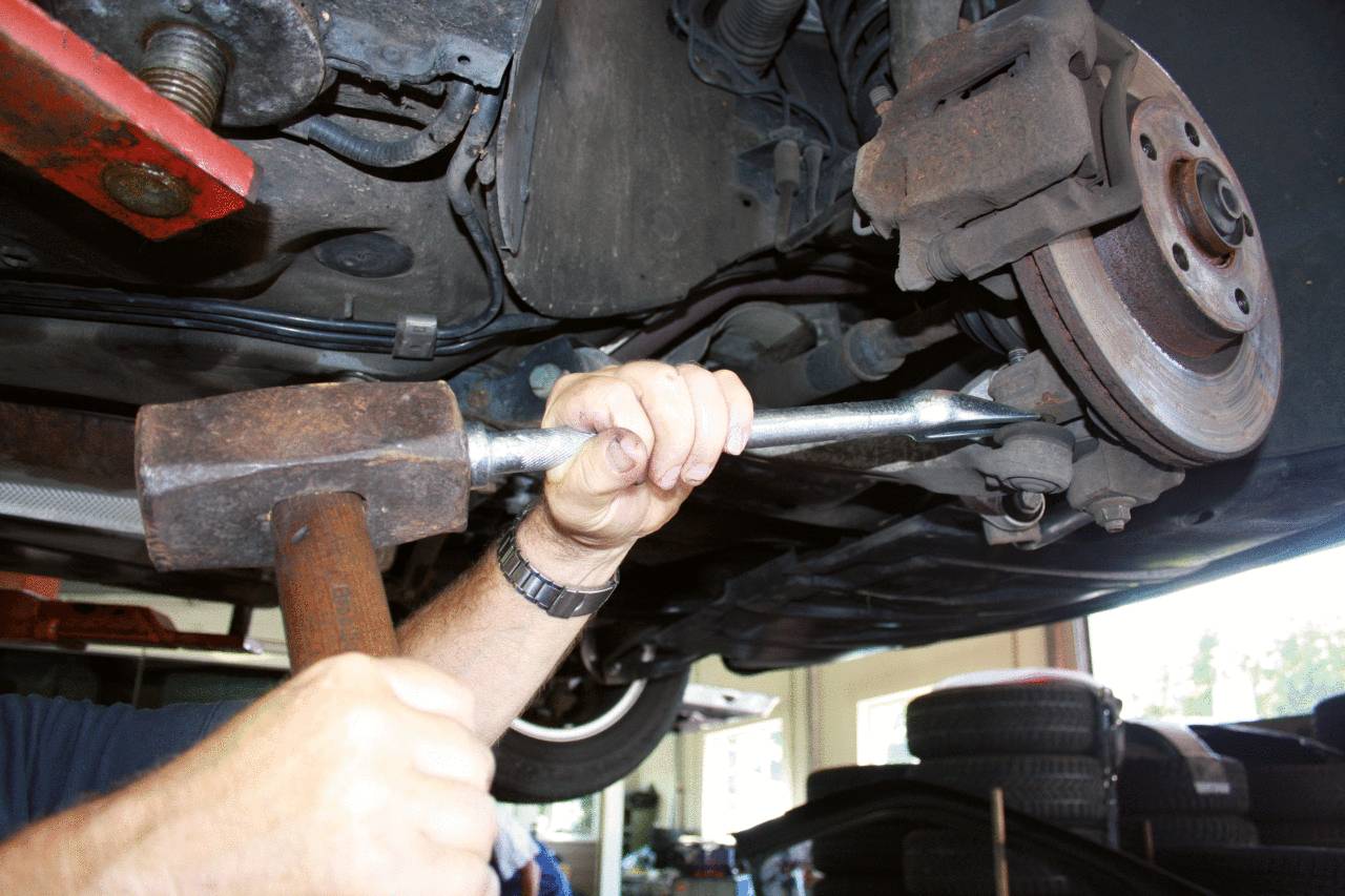 Eine Person repariert das Fahrwerk eines Autos in einer Werkstatt. Sie benutzt einen großen Hammer und eine Metallstange zur Justierung.