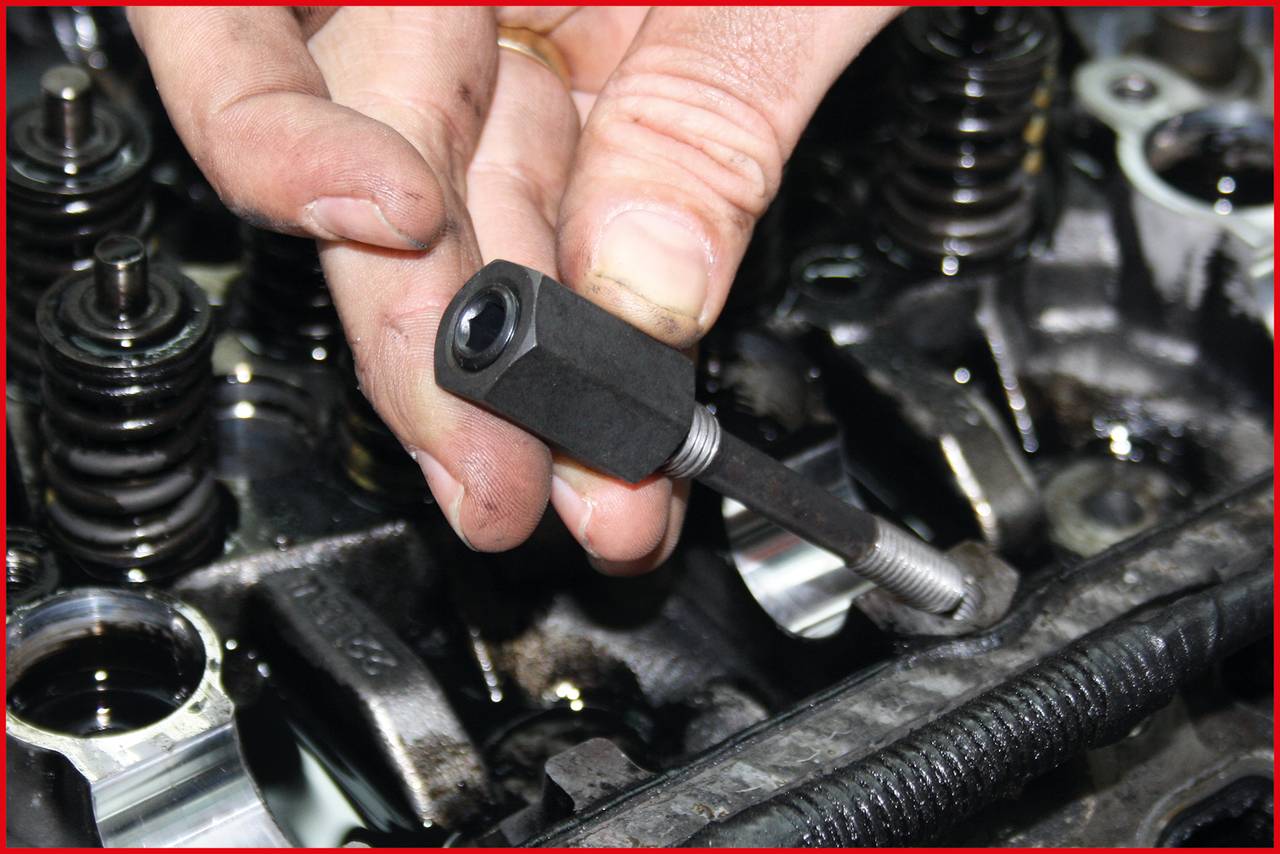 Eine Hand hält einen Schrauben-Extraktor über einem Motorzylinderkopf, umgeben von Ventilfedern und Öl.