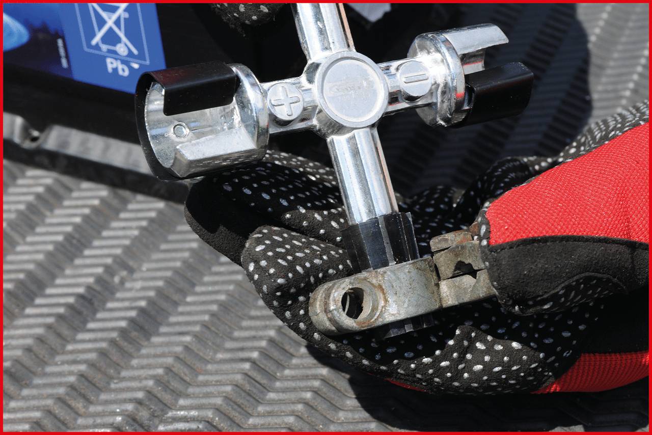 Person mit schwarz-rotem Handschuh hält einen Batterieklemmen-Entferner auf einer metallischen Oberfläche.