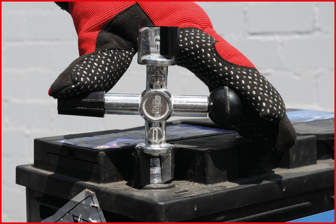 Schwarz-graue Autobatterie mit einer silbernen Überwurfschlüssel, gehalten von Handschuhen, vor einer grauen, gemauerten Wand.