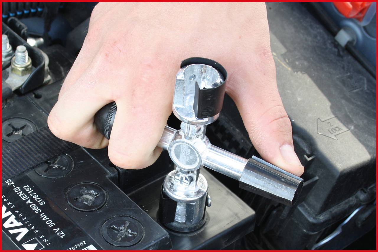 Eine Hand hält ein Werkzeug, um eine Autobatterie zu installieren oder zu entfernen. Die Batterie zeigt Plus- und Minuspol-Markierungen.