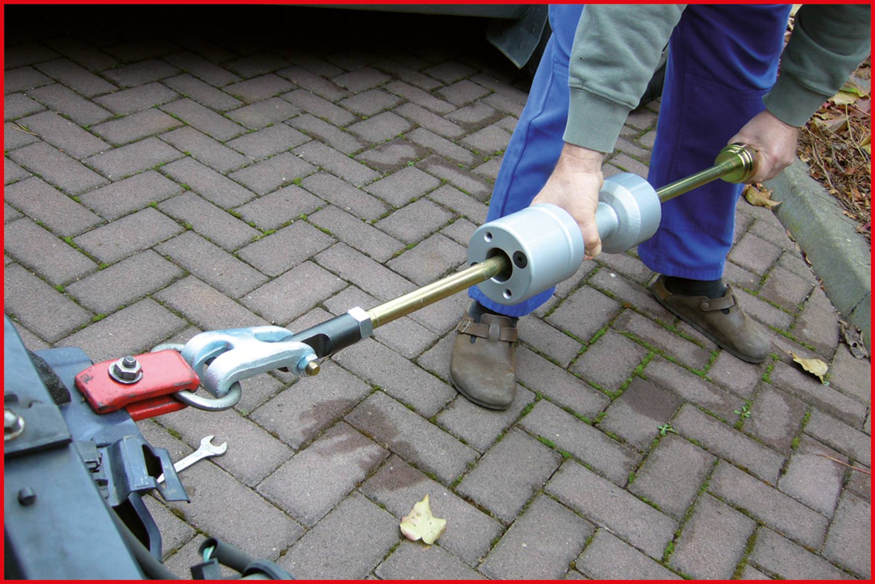 Eine Person repariert die Anhängerkupplung eines Fahrzeugs auf gepflastertem Boden mit einem Werkzeug in der Hand.