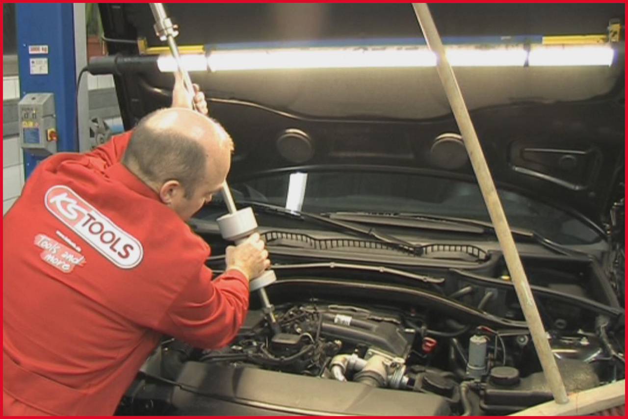 Ein Mechaniker arbeitet mit einem Werkzeug unter der geöffneten Motorhaube eines Autos in einer Werkstatt.