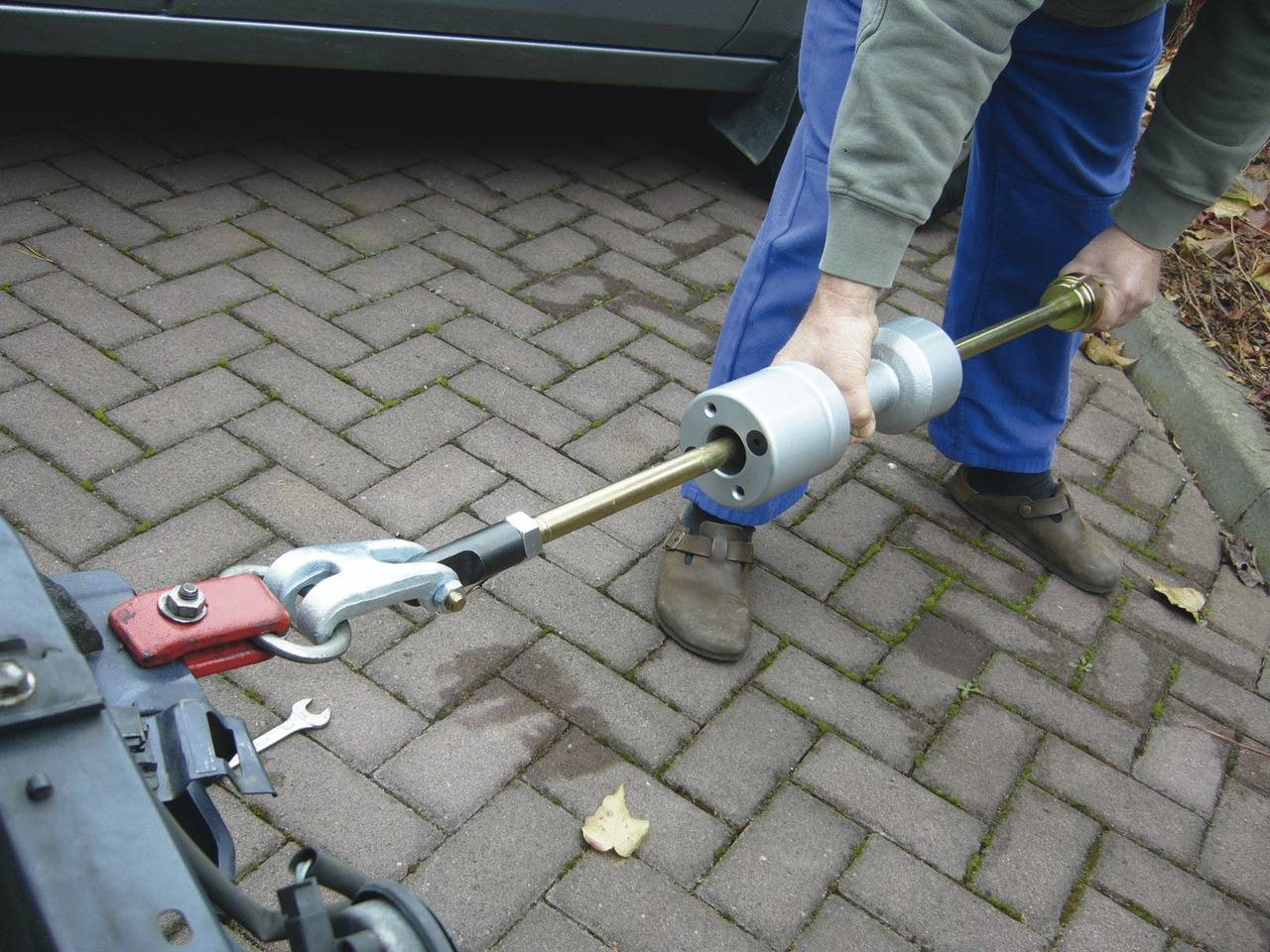 Person befestigt eine Stange an einer Anhängerkupplung mit einem Werkzeug. Autoreifen und Pflastersteine sichtbar im Hintergrund.