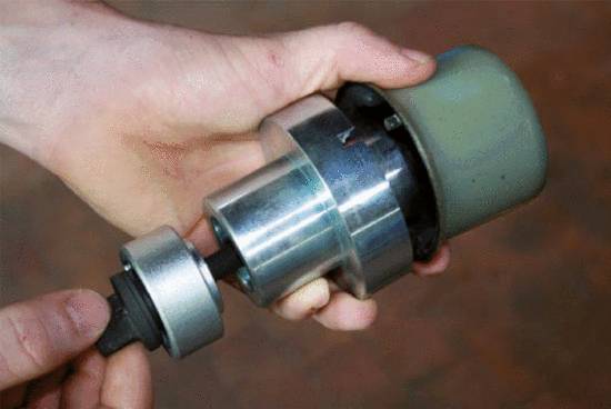Hände halten einen metallischen Zylinder mit einem abnehmbaren Teil. Es scheint sich um eine mechanische Komponente zu handeln.