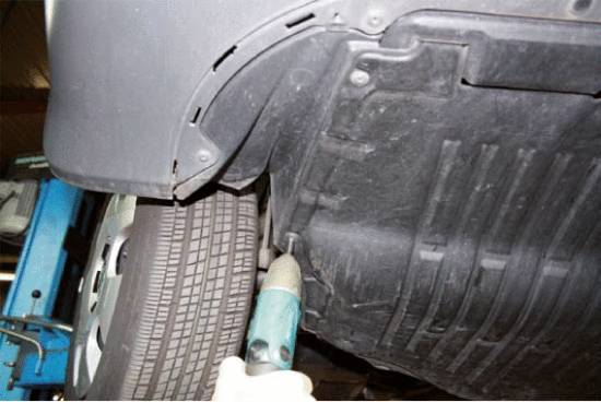 Mechaniker führt Reparatur an der Unterseite eines aufgebockten Autos durch. Er benutzt ein elektrisches Werkzeug nahe dem Hinterrad.