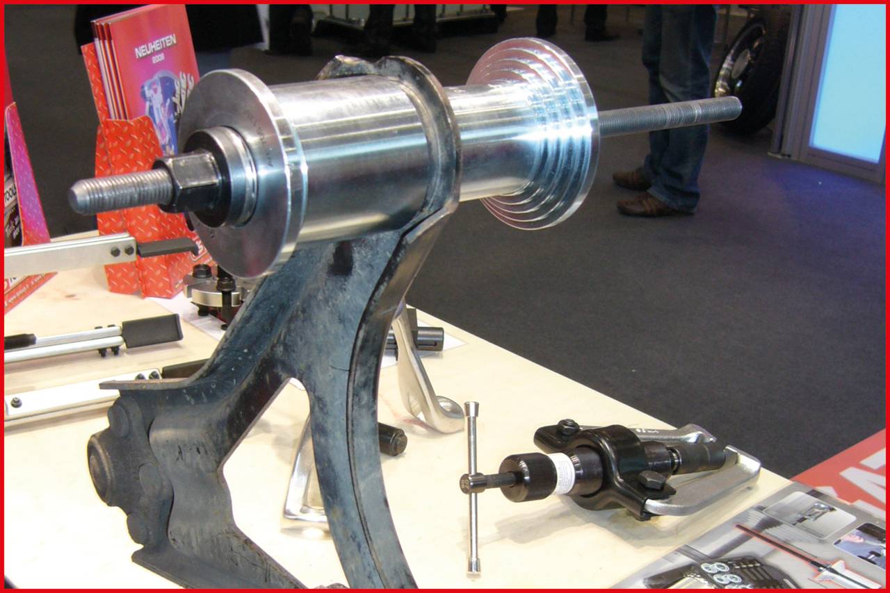 Maschinenteile auf einem Tisch ausgestellt, darunter eine Metallwelle in einem Halter, umgeben von Werkzeugen und Prospekten.