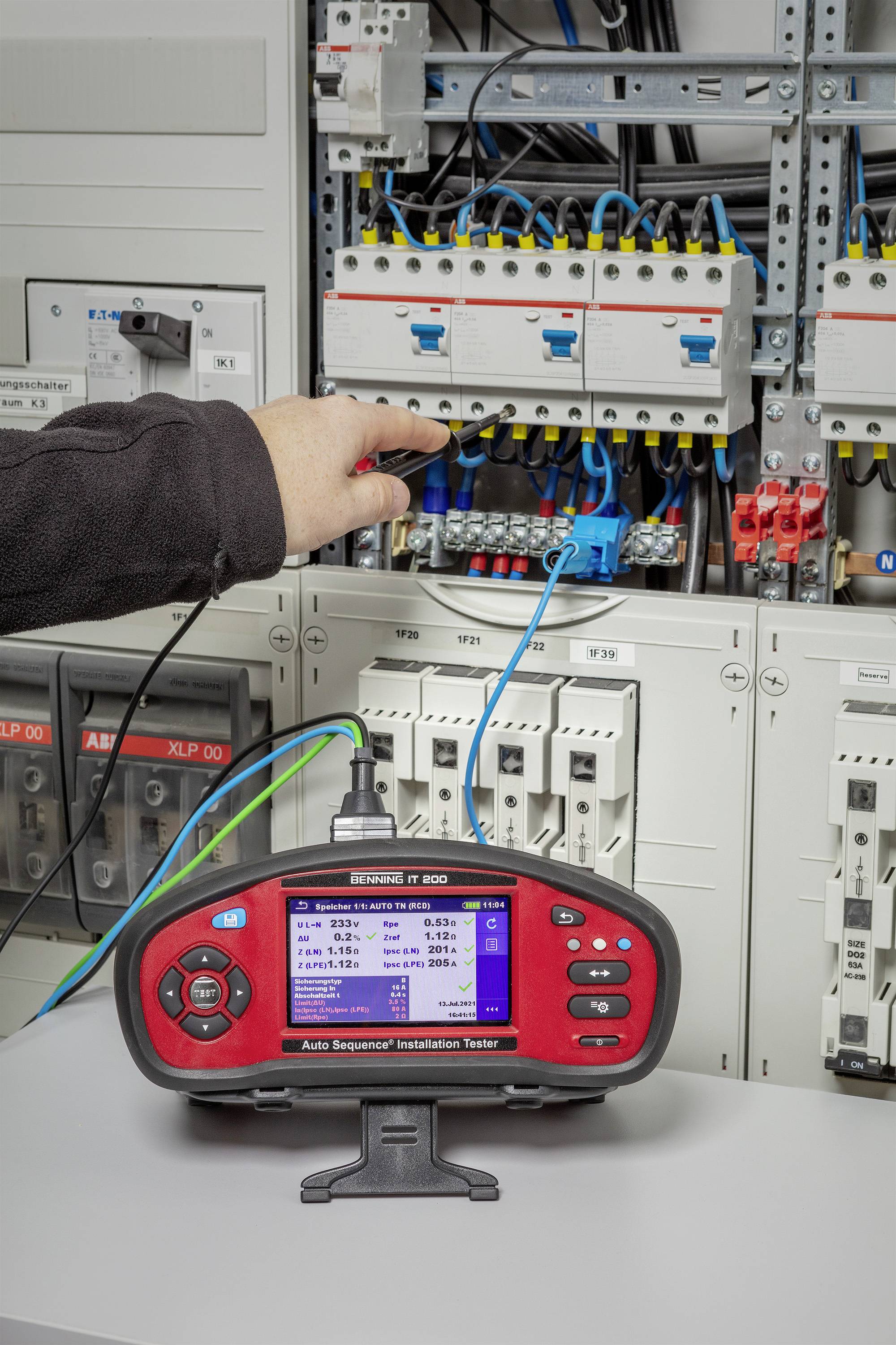 Ein Techniker misst mit einem Elektroprüfgerät den Strom in einem Schaltschrank. Verschiedene Kabel und Sicherungen sind sichtbar.