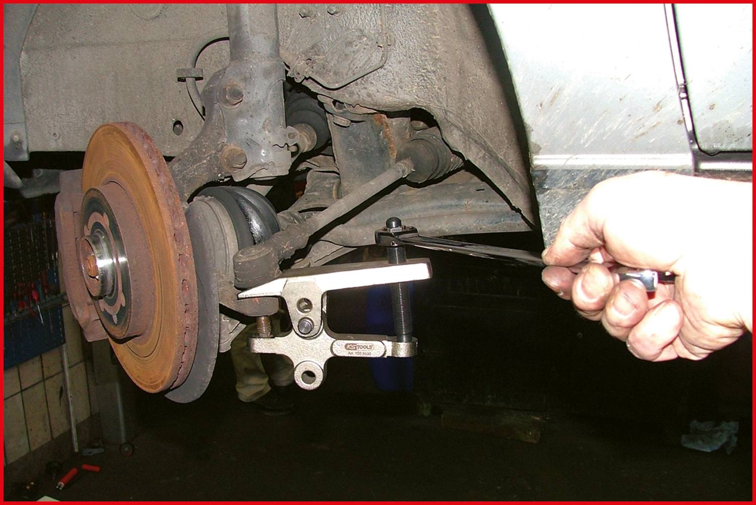 Ein Mechaniker benutzt ein Ziehwerkzeug, um das Kugelgelenk von einem Fahrzeug zu entfernen. Im Hintergrund ist ein Bremsscheibenrotor zu sehen.
