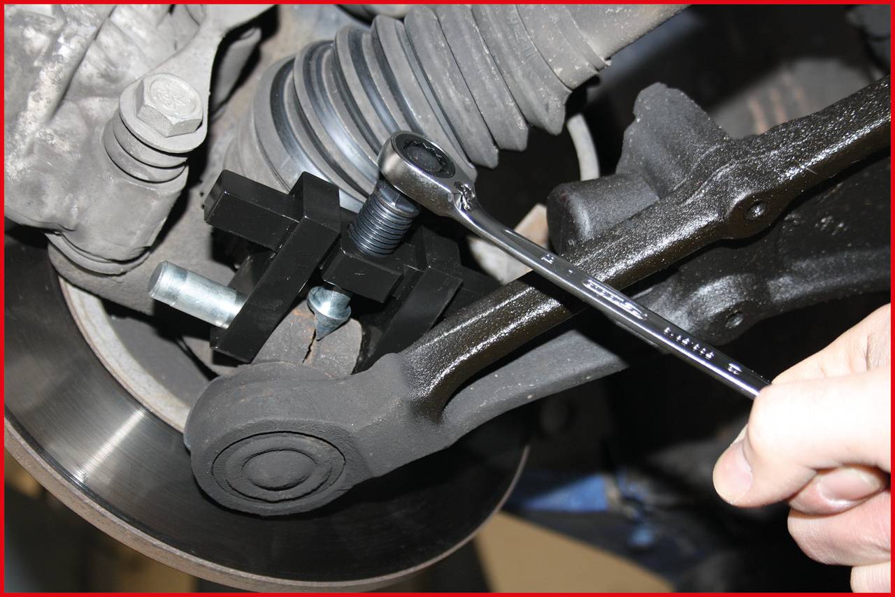 Eine Hand zieht mit einem Schraubenschlüssel eine Schraube an der Achse eines Autos fest. Die Aufhängung und Bremsscheibe sind sichtbar.