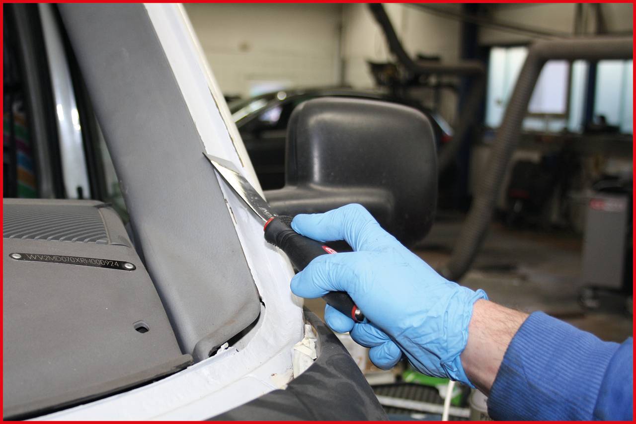 Person mit blauen Handschuhen schneidet mit einem Werkzeug die Dichtung eines Autofensters in einer Werkstatt.