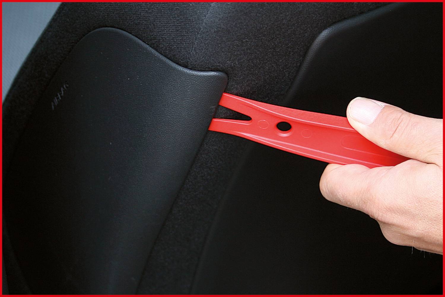 Eine Hand hält ein rotes Hebelwerkzeug, das zwischen einem schwarzen Autoinnenraum und einem Bauteil eingesetzt wird.