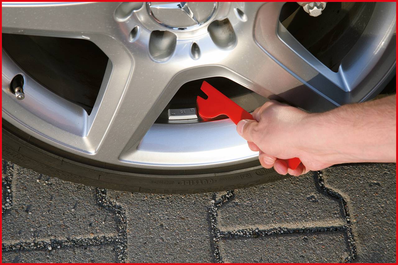 Eine Hand hält ein rotes Werkzeug und misst den Abstand zwischen einer Autospeiche und der Bremsscheibe an einem silbernen Autoreifen.