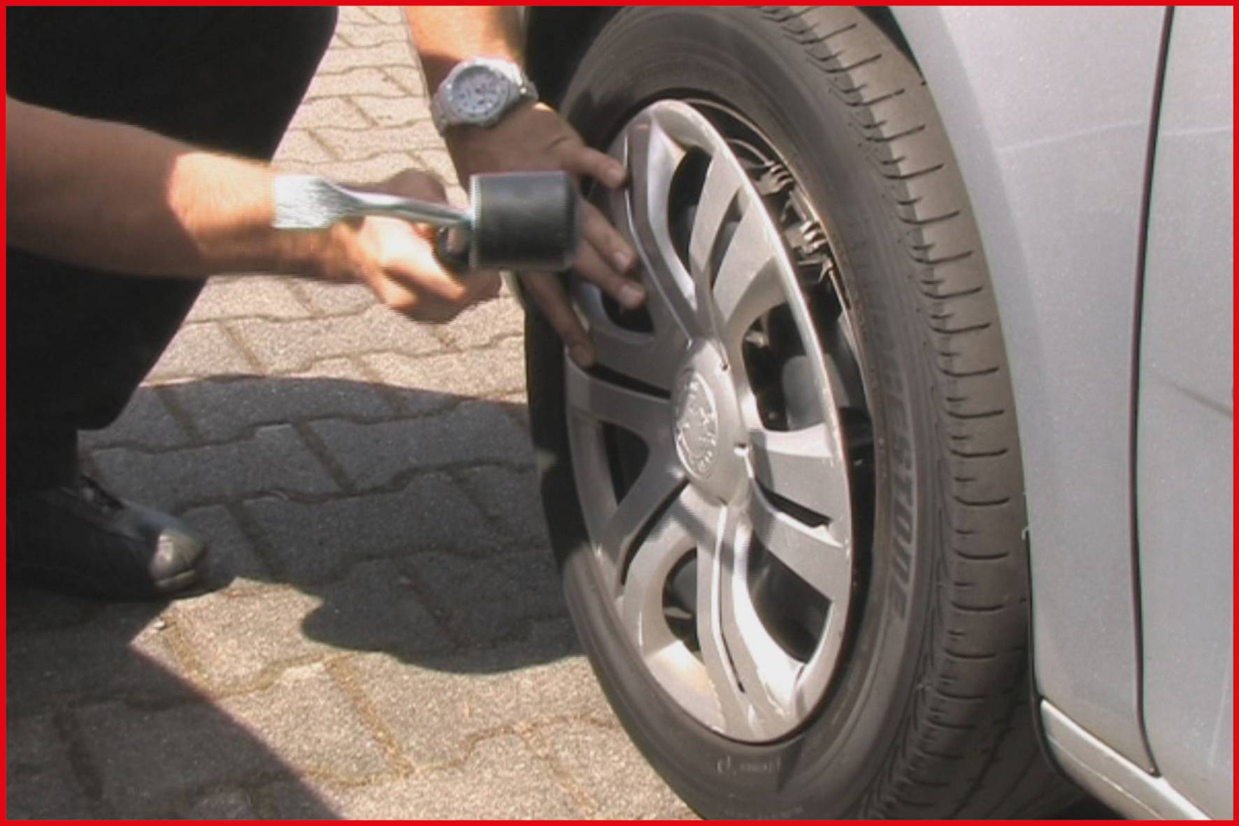 Eine Person repariert oder prüft ein Autoreifen mit einem Werkzeug. Der Reifen ist an einem grau-silbernen Auto montiert.