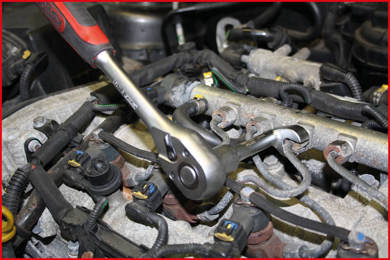 Ein Ratschenwerkzeug wird an einem Fahrzeugmotor verwendet, um an der Zündkerze zu arbeiten. Verschiedene Kabel und Anschlüsse sind sichtbar.