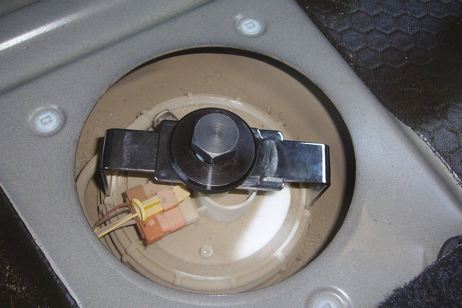 'Runder Deckel im Auto öffnet Zugang zur Kraftstoffpumpe. Oben ist ein Verschluss mit einem Bolzen, daneben verlaufen Kabel.'