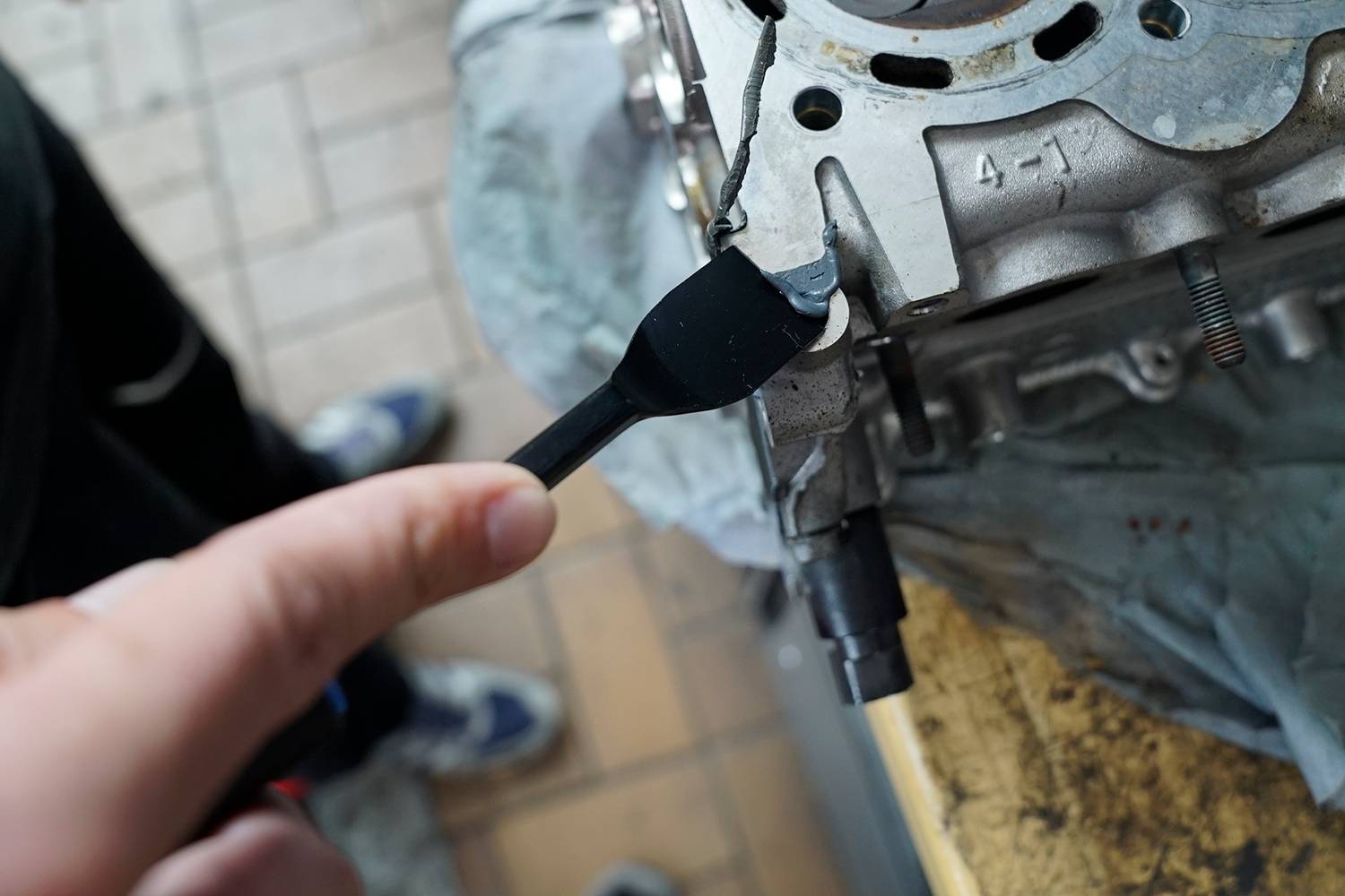 Eine Person trägt eine Schicht Dichtmasse auf die Kante eines Metallmotors auf. Der Motor liegt auf einem Tisch.