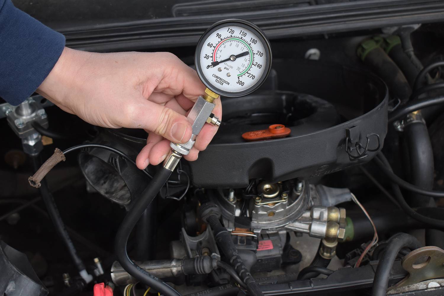 Eine Person hält ein Manometer über einem Autoteil im Motorraum, um den Druck zu prüfen. Das Manometer zeigt Messwerte in zwei Skalen an.