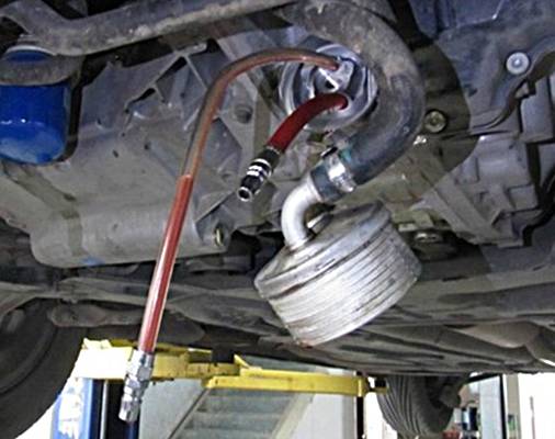 'Ölablassvorgang bei einem Fahrzeug in einer Werkstatt. Ein Schlauch leitet das abgelassene Öl von der Unterseite des Motors in einen Behälter.'