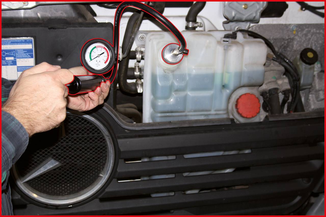 Eine Person prüft den Druck in einem Auto-Kühlsystem. Ein Manometer ist an einen Behälter angeschlossen, der teilweise mit blauem Kühlmittel gefüllt ist.