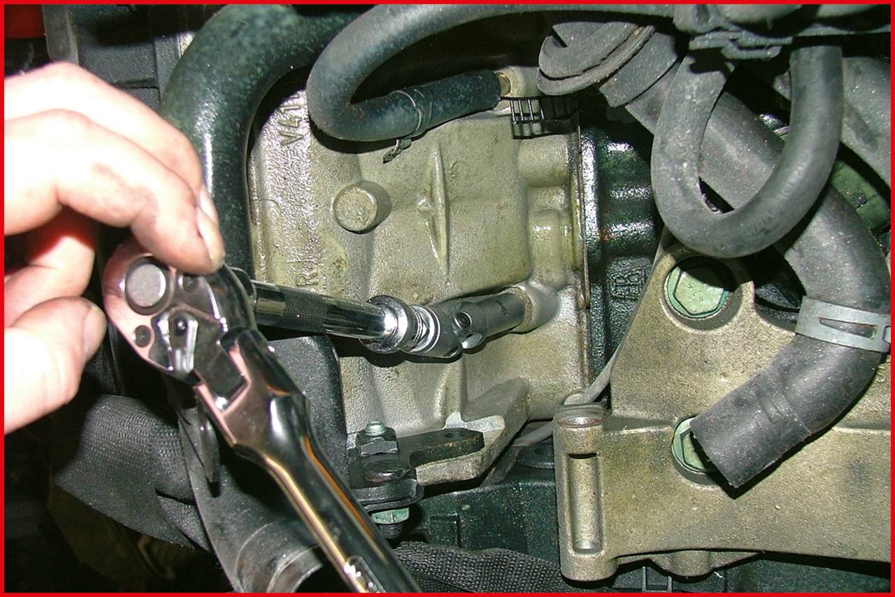 Eine Person zieht mit einem Ratschenwerkzeug eine Schraube im Motorbereich eines Fahrzeugs an. Schläuche und Teile sind um den Arbeitsbereich sichtbar.