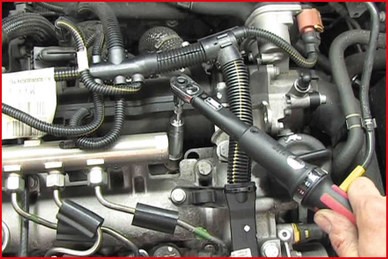 Eine Hand hält ein Ratschenschlüssel, der in einem Auto-Motorraum verwendet wird, um an einem Bauteil zu arbeiten. Kabel und Rohre sind sichtbar.