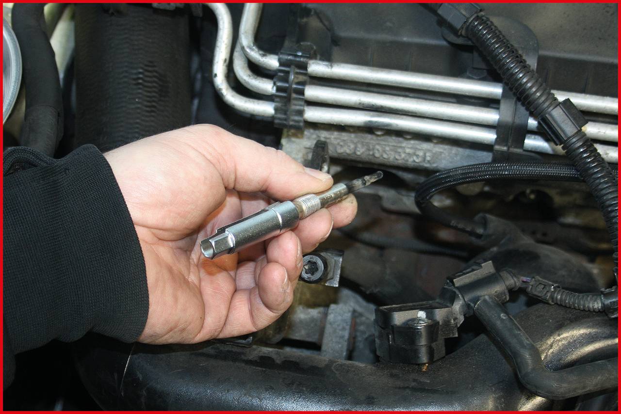 Eine Hand hält eine Glühkerze vor einem Motorblock. Mechanische Teile und Kabel sind im Hintergrund sichtbar.