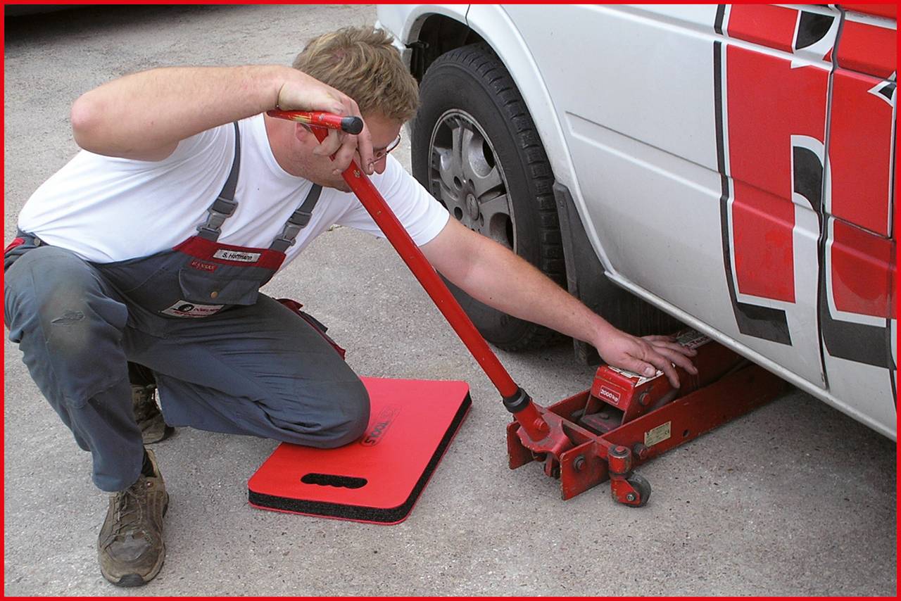 Ein Mann wechselt einen Reifen, verwendet einen Wagenheber, um ein Auto anzuheben. Er trägt Arbeitskleidung und kniet auf dem Boden.