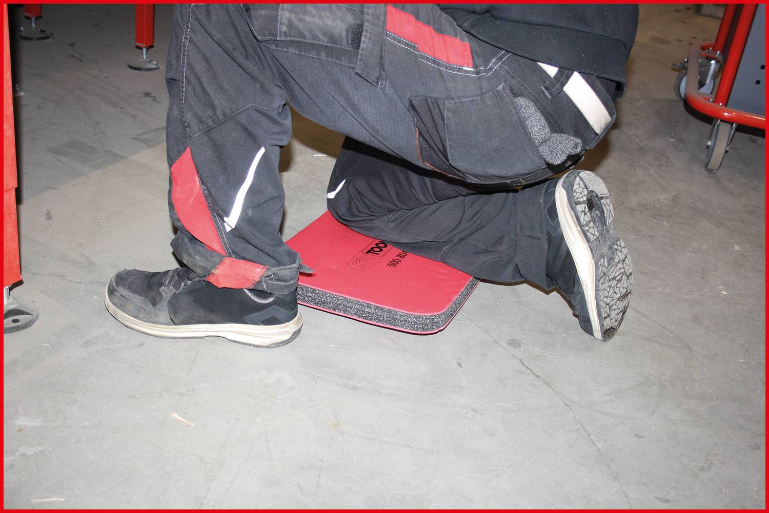 Eine Person kniet auf einer roten Matte auf einem Betonboden und trägt schwarze Arbeitsschuhe und eine schwarze Hose mit roten Streifen.
