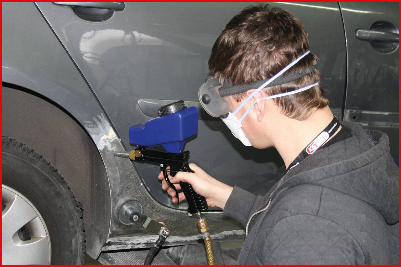 Ein Mann trägt Schutzbrille und Mundschutz, während er mit einer Sprühpistole einen Autoteil lackiert.