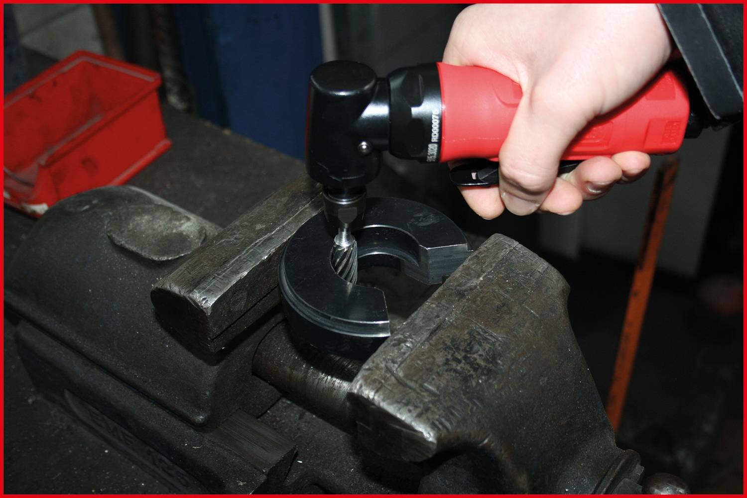 'Eine Hand hält einen roten elektrischen Werkzeugschrauber, der auf einem Metallstück in einem Schraubstock arbeitet.'