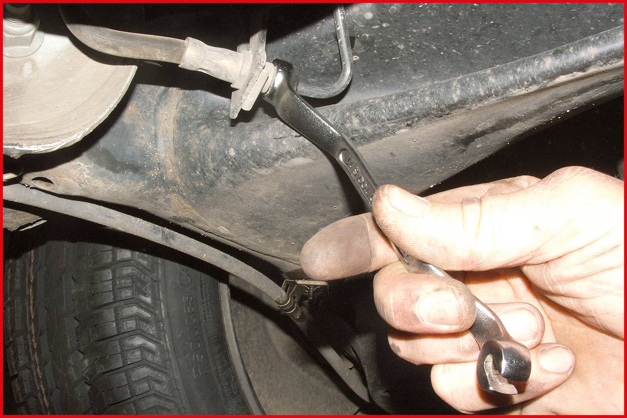 Eine Hand hält einen Schraubenschlüssel, um einen Fahrzeugbremsmechanismus zu reparieren. Ein Reifen und Unterbodenteile sind sichtbar.