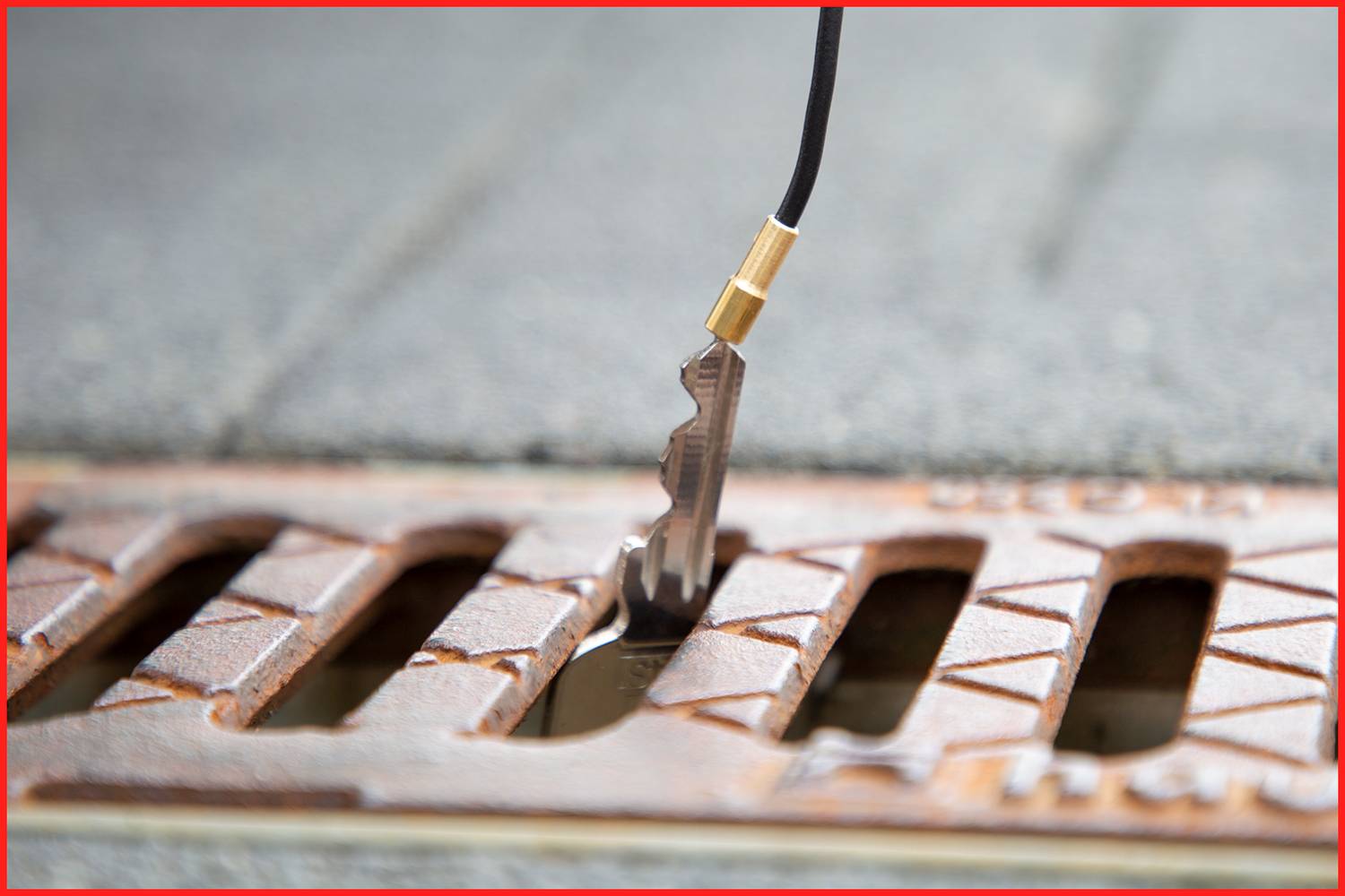 Ein Metallobjekt, das wie ein Schlüssel aussieht, steckt in einem Abflussgitter auf einer Straße. Ein Kabel ist mit dem Objekt verbunden.