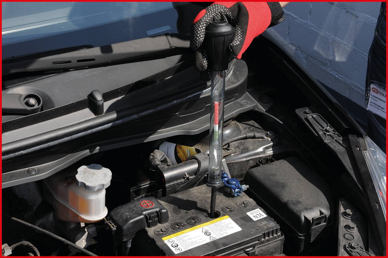 Person mit roten Handschuhen testet den Ladezustand einer Autobatterie unter der geöffneten Motorhaube mit einem Batterieprüfgerät.