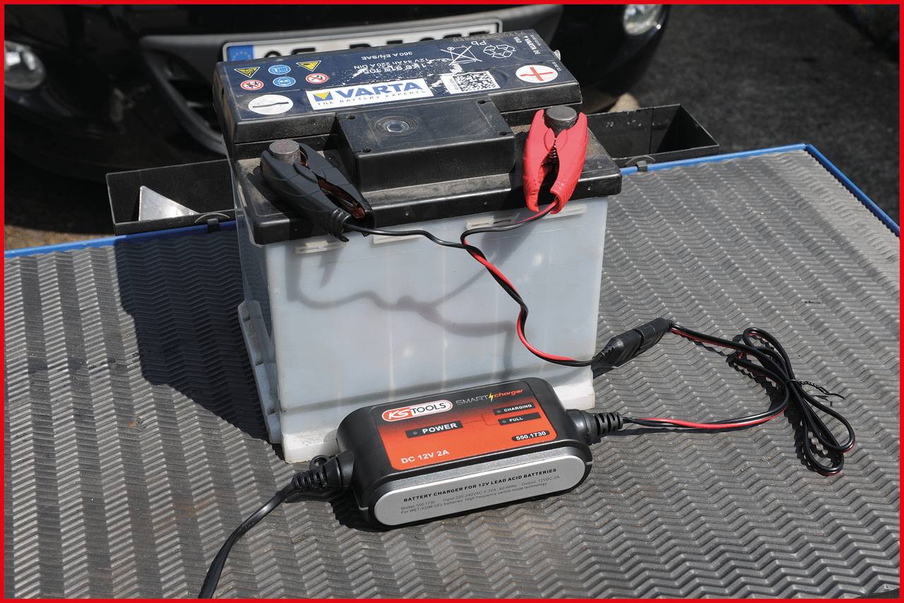 Auto-Batterie auf einem Tisch mit angeschlossenem Ladegerät. Ladegerät hat rote und schwarze Kabel, die an die Batterie geklemmt sind.