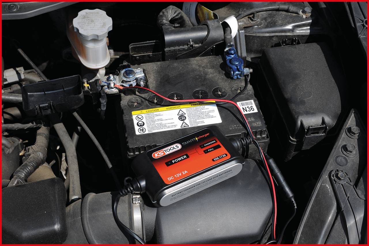 Batterieladegerät, das an eine Autobatterie angeschlossen ist. Das Gerät zeigt Betriebsanzeige-LEDs. Motorraum im Hintergrund.