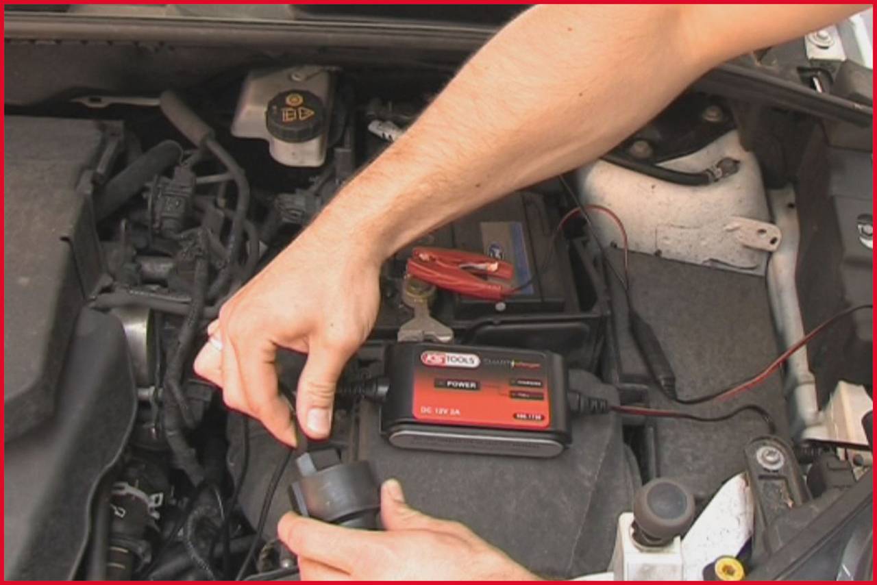 Eine Person prüft die Autobatterie mit einem Diagnosetool im Motorraum, hält einen Adapter in der einen Hand.