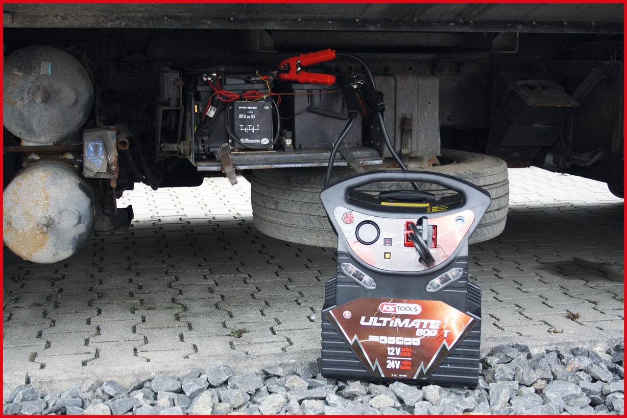 'Ein Batterieladegerät ist an eine Autobatterie angeschlossen, um sie aufzuladen. Der Lkw steht auf einem gepflasterten Untergrund.'