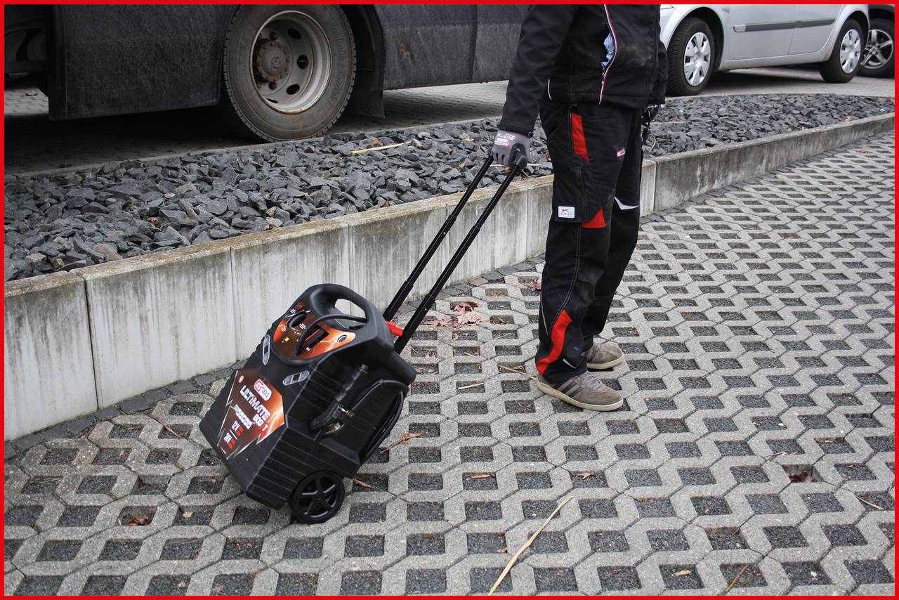 Eine Person zieht einen schwarzen Wagen mit einem Griff über einen gepflasterten Gehweg. Im Hintergrund sind Fahrzeuge zu sehen.