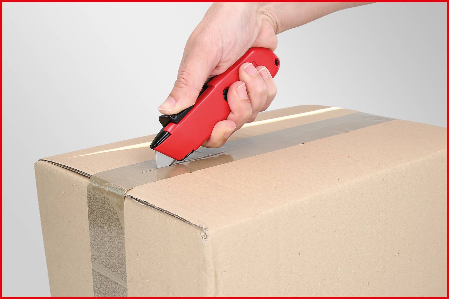 Hand mit rotem Messer schneidet Klebeband auf einem braunen Karton auf.