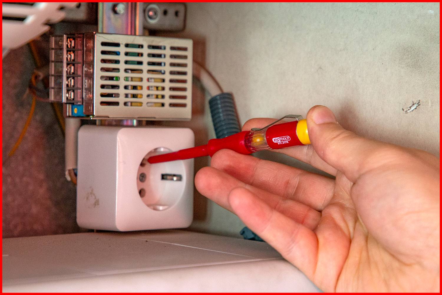 Eine Hand hält einen spannungsprüfenden Schraubendreher an einer weißen Steckdose, um die Spannung zu messen.