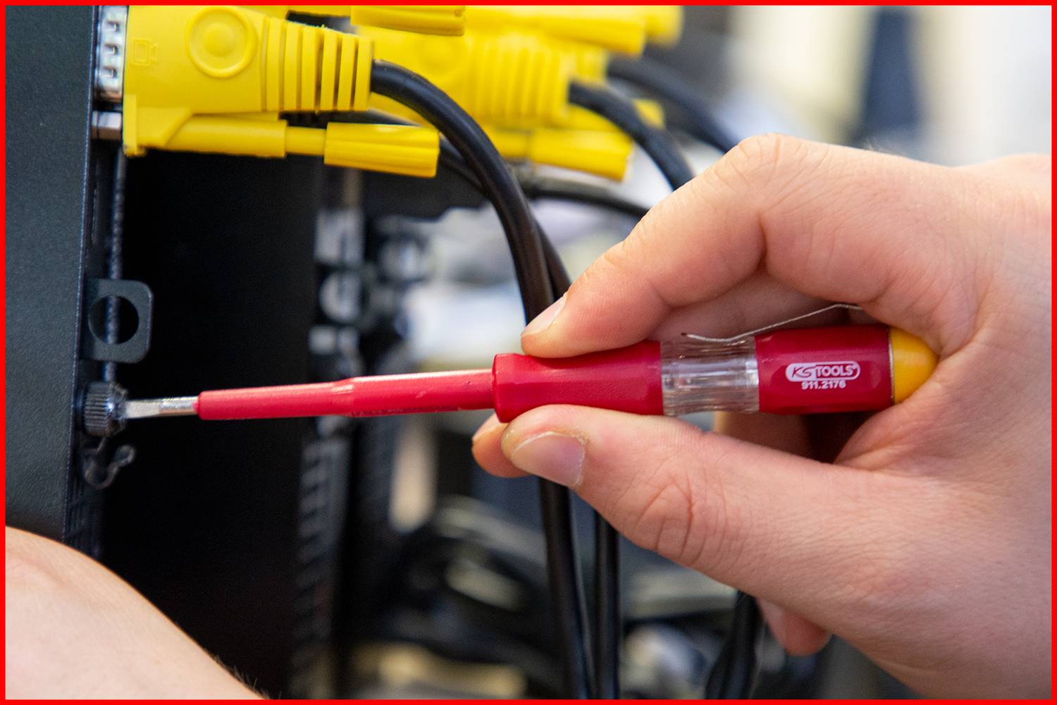 Eine Hand schraubt mit einem roten Schraubendreher ein Kabel in ein elektronisches Gerät. Mehrere gelbe Kabel sind eingesteckt.