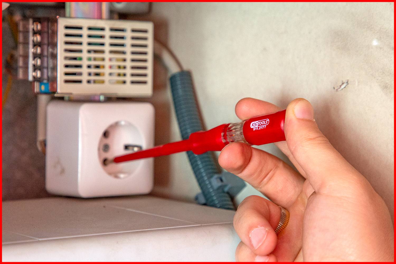 Eine Hand testet mit einem Spannungsprüfer eine Steckdose. Ein blaues Kabel ist sichtbar. Sicherheitsprüfungssituation.
