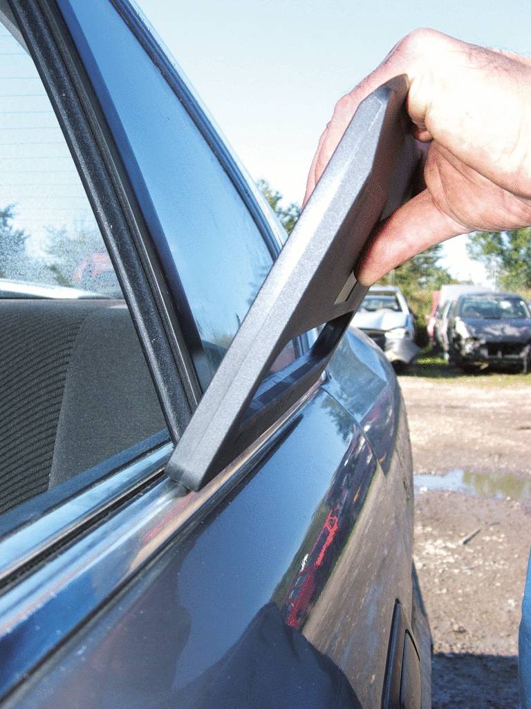 Eine Hand öffnet mit einem Keil ein Autofenster, vermutlich zur Autoöffnung oder Pannenhilfe. Im Hintergrund stehen weitere Autos.